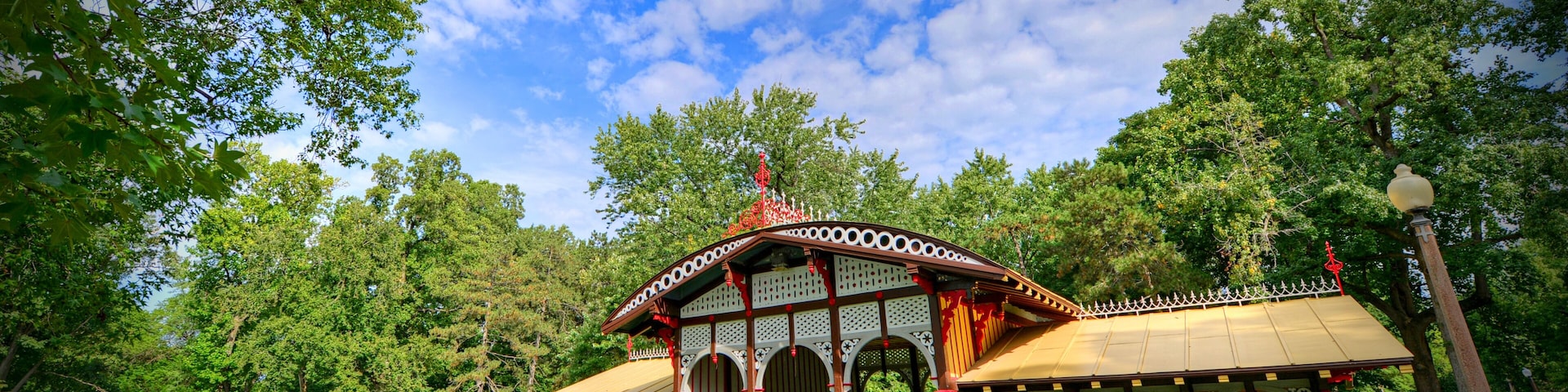 Um gazebo com telhado vermelho e paredes brancas, cercado por árvores e grama verdejantes.