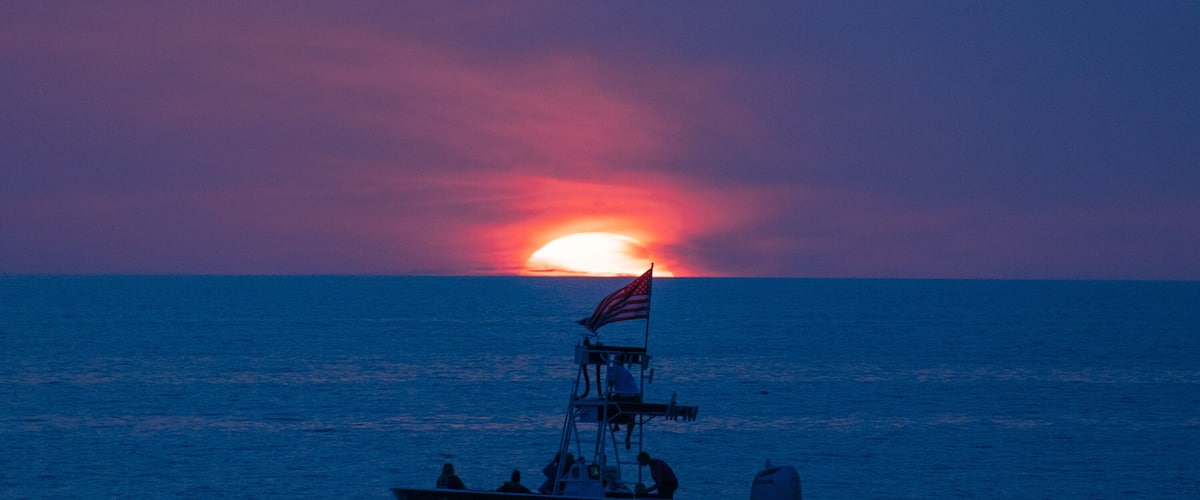 Closeup of a boat on the ocean in front of a glowing setting sun amid the blue and purple clouds of evening, as seen from a beach on the Gulf of Mexico near Englewood, Florida, USA, in early spring