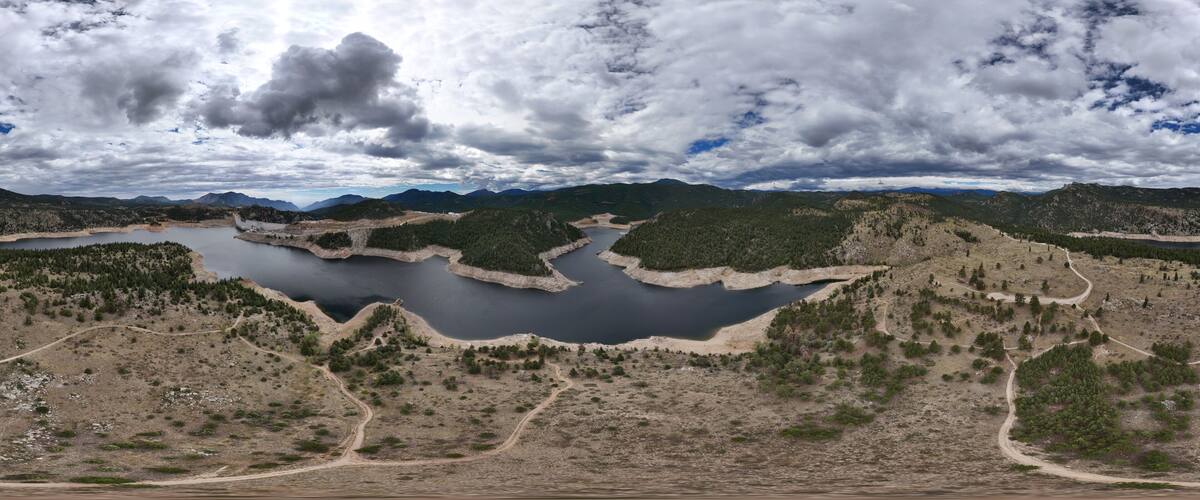 Aerial view of Gross Reservoir surrounded by hills, pine trees, and dirt trails under a cloudy sky in Boulder County, Colorado, United States.