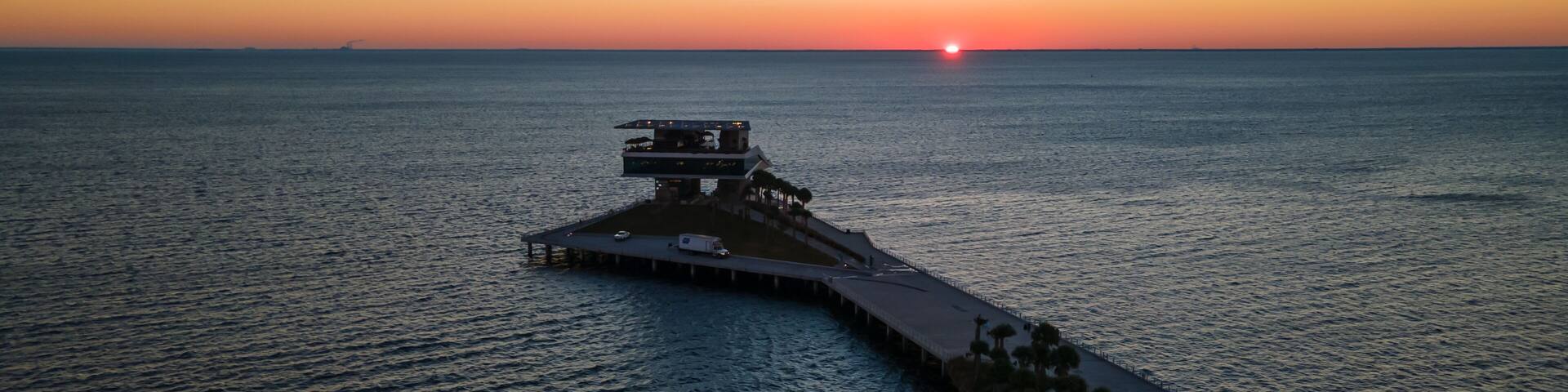 A pier extending into the water with a building on top, palm trees along the side, and the sun setting in the background.