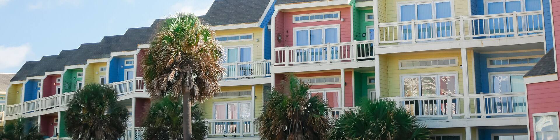 Panorama view beach townhouses framed by palm trees under sunny cloud blue skies in Galveston, Texas. Tropical accents and pastel tones coastal living, open white balcony, upscale rental units