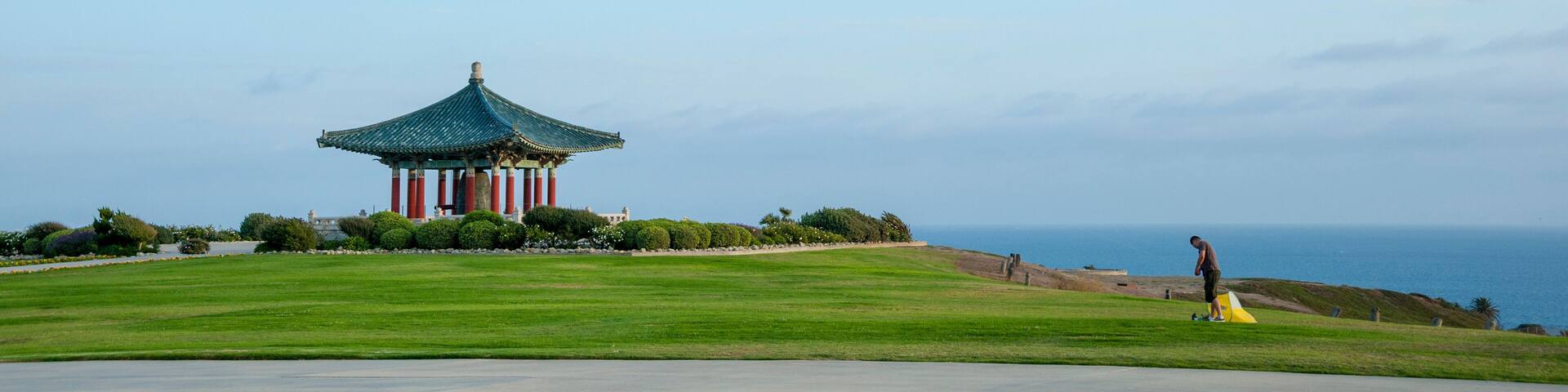 A traditional pavilion with a green roof, a grassy field, and a person flying a kite near the ocean.
