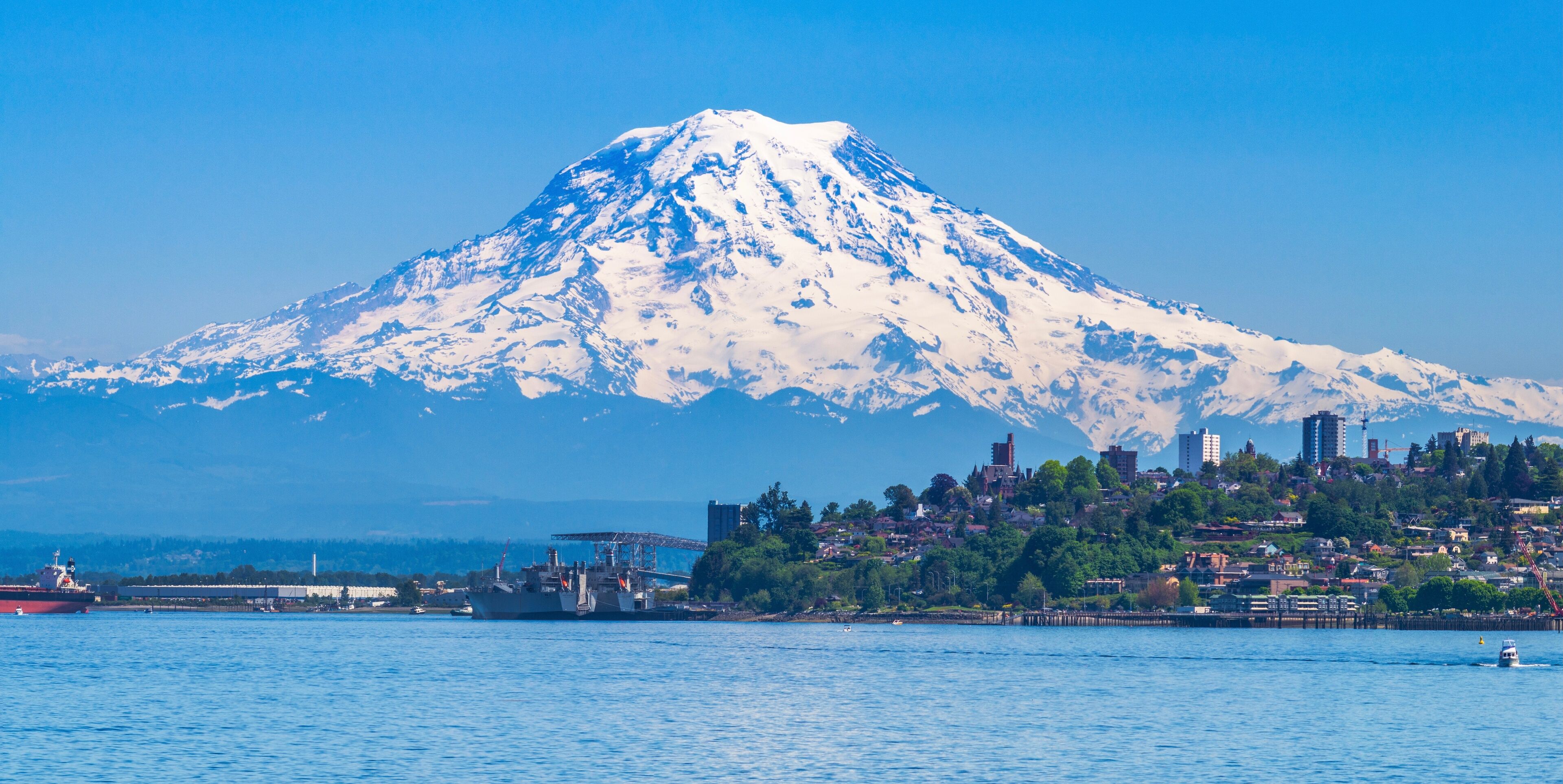 A snow-capped mountain, a body of water, and a cityscape with buildings and greenery.