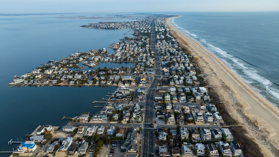 An aerial view of a coastal residential area with a long beachfront, a marina, and a densely packed neighborhood.