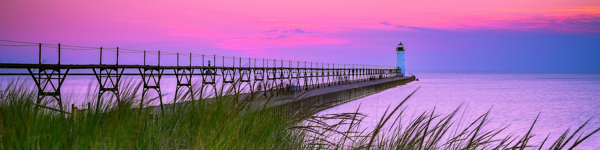 A pier extending into a body of water with a lighthouse at the end, surrounded by tall grass.