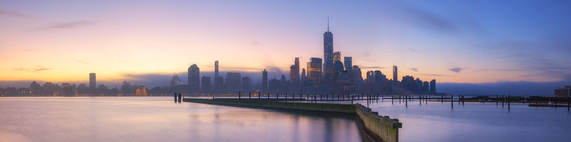 A city skyline at dusk with a calm river in the foreground.