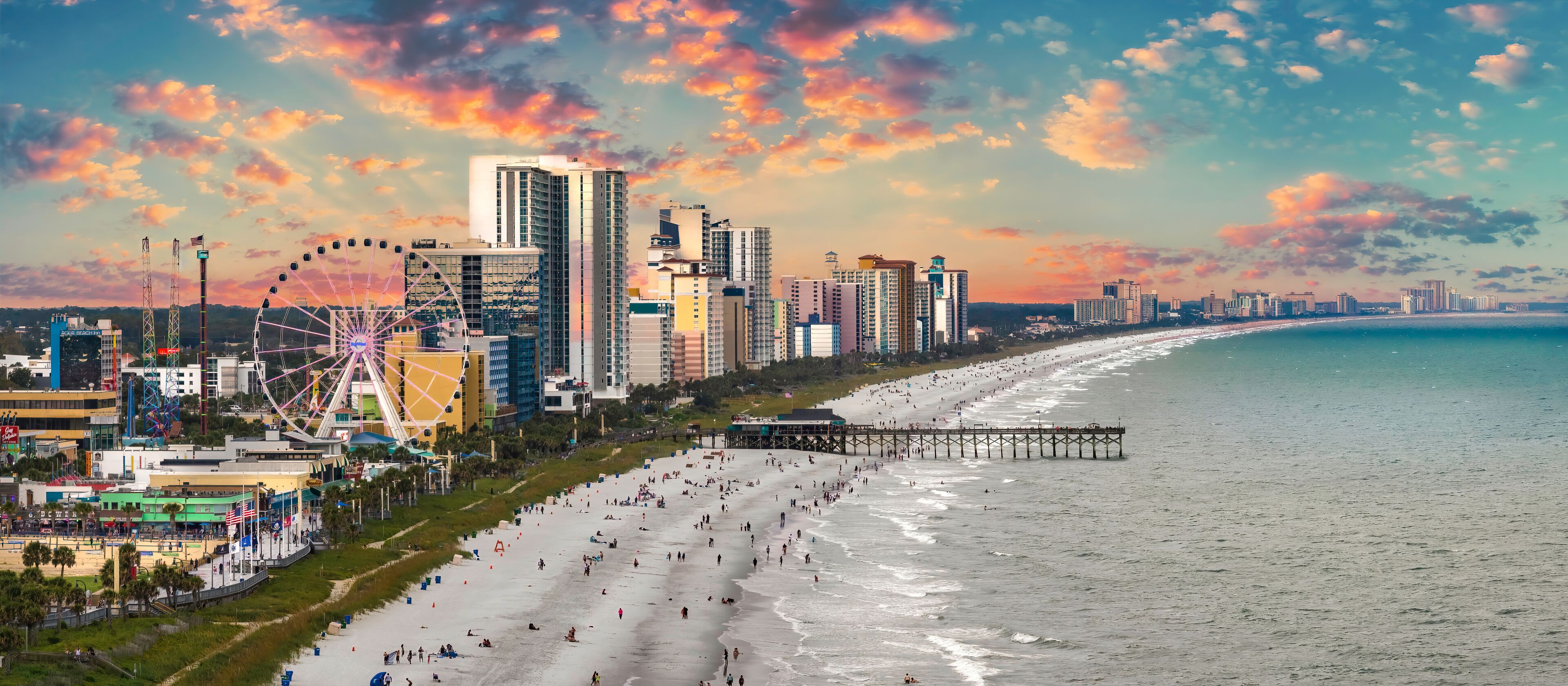 A beach with a pier, a Ferris wheel, and high-rise buildings under a colorful sunset sky.