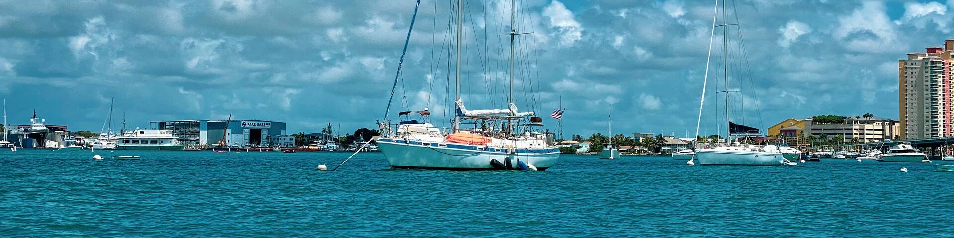 A marina with sailboats and yachts docked, buildings, and a clear blue sky with clouds.