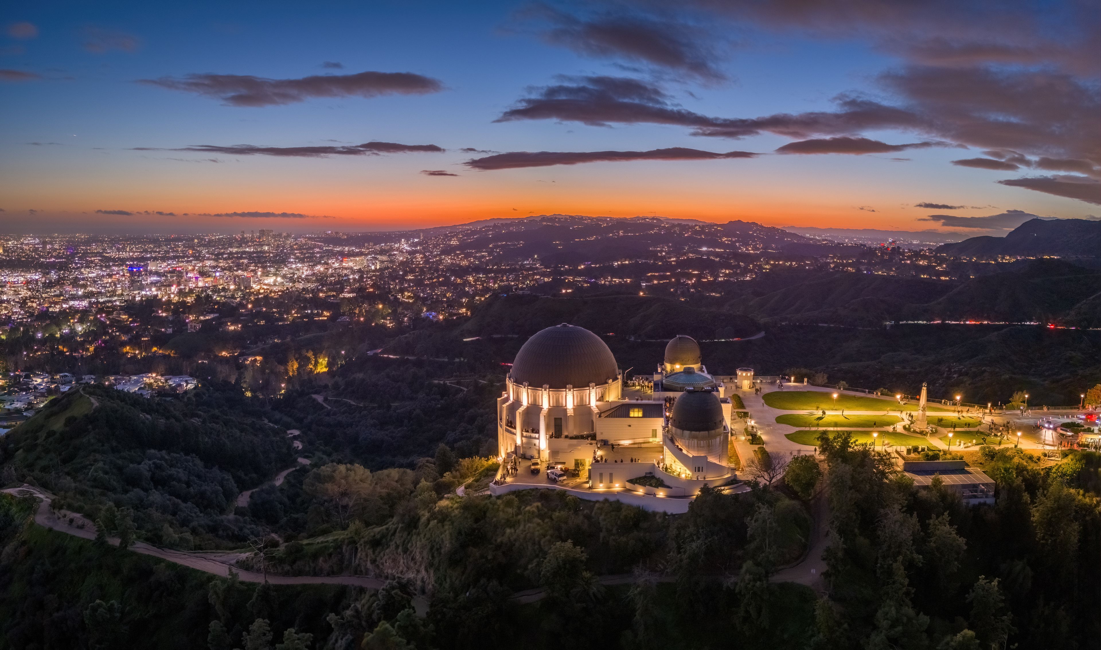 A cityscape at dusk with two prominent domed buildings, a winding road, and a hilly landscape.