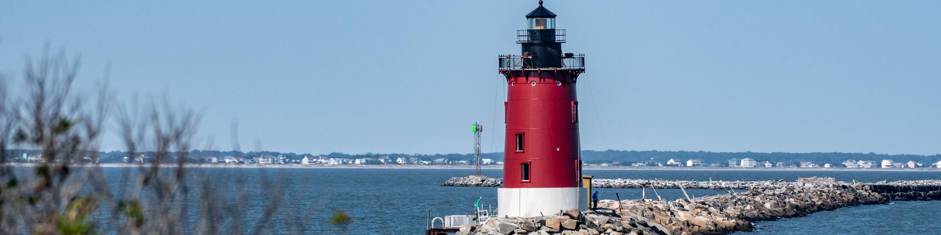 A red and white lighthouse on a rocky outcrop with calm water in the foreground and a clear sky.