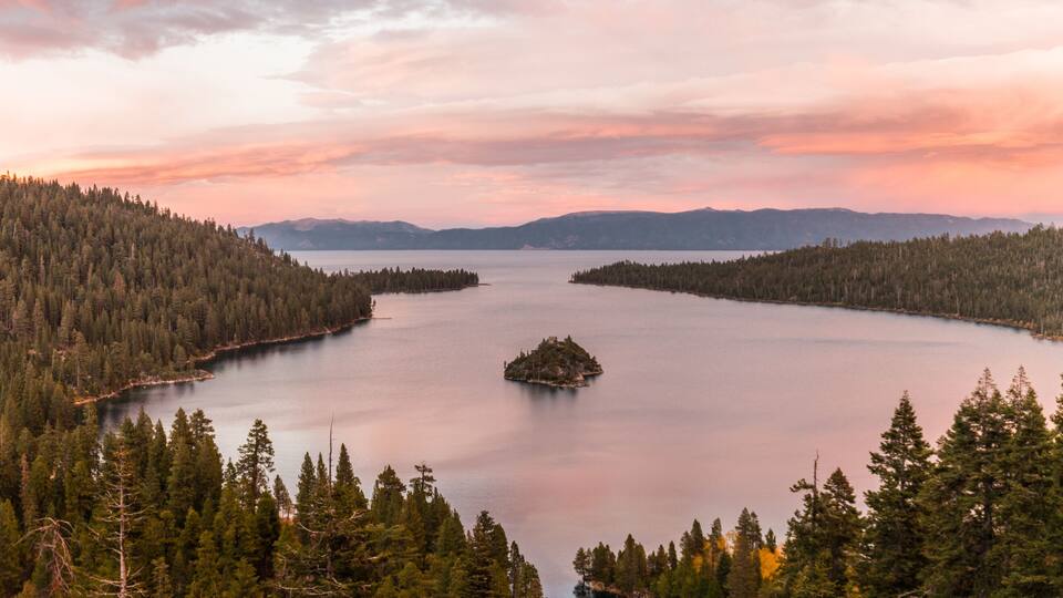 Un lac entouré de forêts denses et de montagnes, sous un ciel rose.