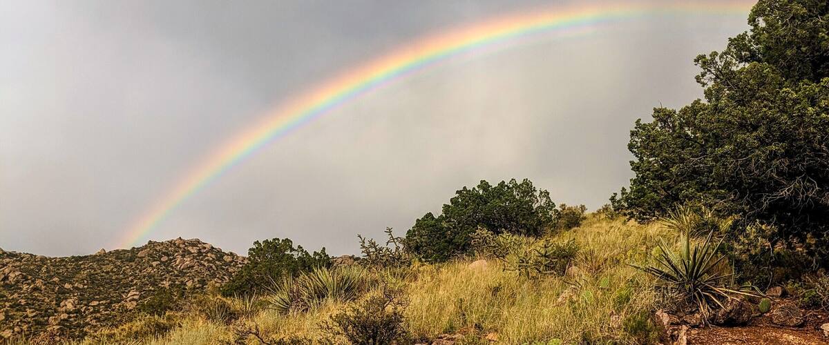 A landscape with a rainbow arching over a rocky terrain with sparse vegetation.