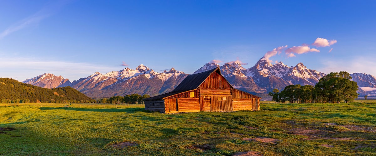 A wooden cabin in a grassy field with snow-capped mountains in the background.