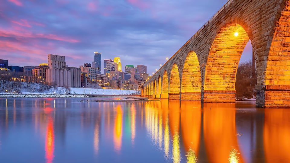 A stone bridge over a calm river reflecting city lights, with a snow-covered area and modern buildings in the background.