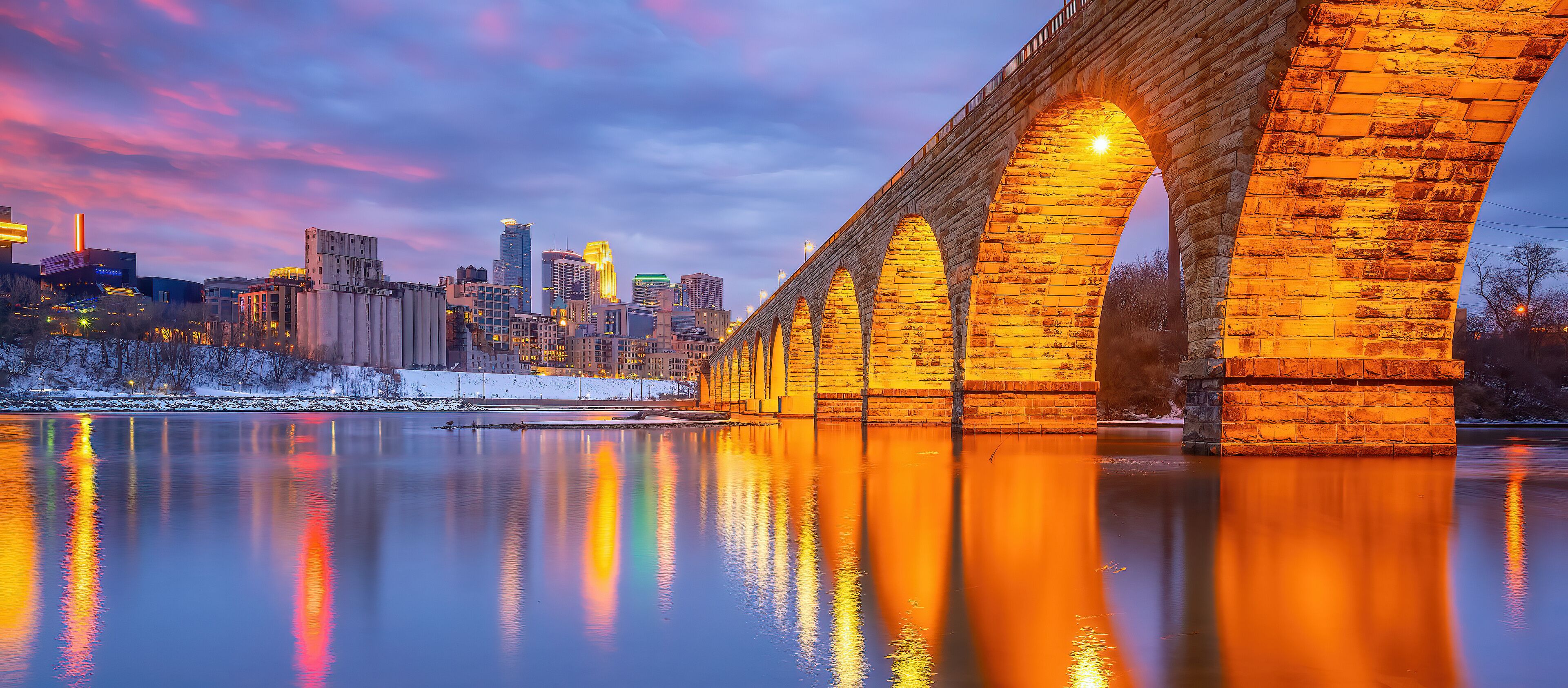 A stone bridge over a calm river reflecting city lights, with a snow-covered area and modern buildings in the background.