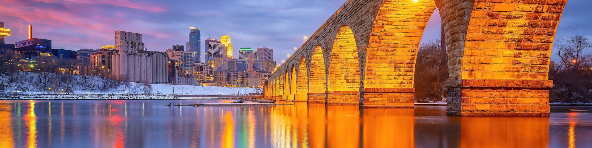 A stone bridge over a calm river reflecting city lights, with a snow-covered area and modern buildings in the background.