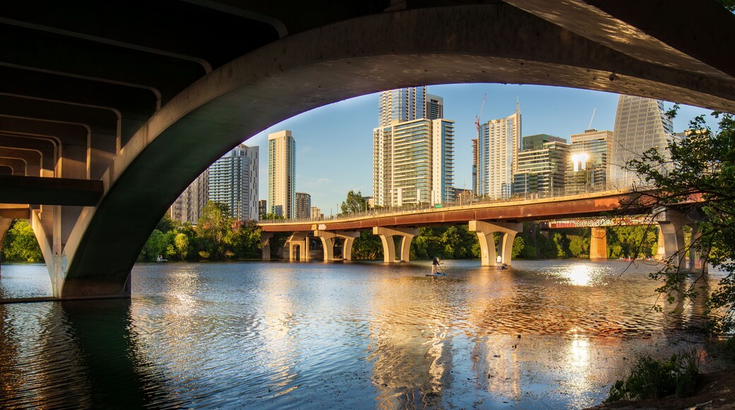 Beneath the North Lamar Bridge in Austin, Texas, the tranquil waters of Lady Bird Lake mirror the golden hues of the cityscape, creating a beautiful reflection on a summer day.