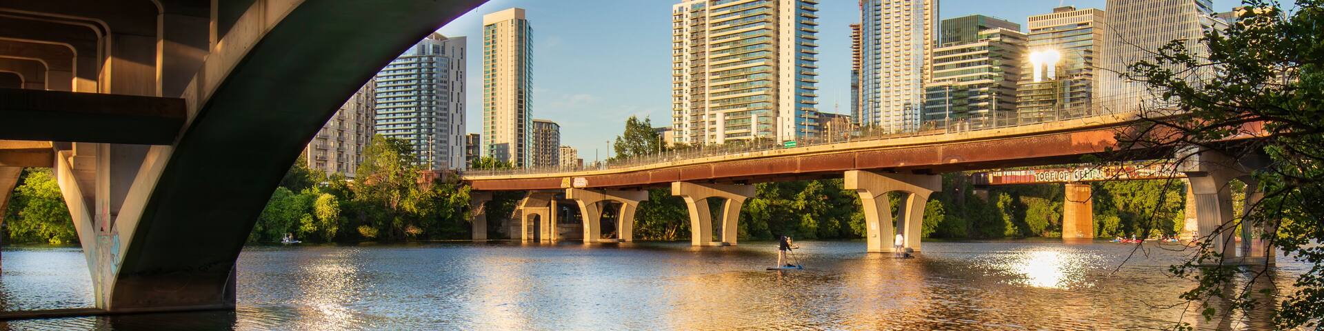 Beneath the North Lamar Bridge in Austin, Texas, the tranquil waters of Lady Bird Lake mirror the golden hues of the cityscape, creating a beautiful reflection on a summer day.