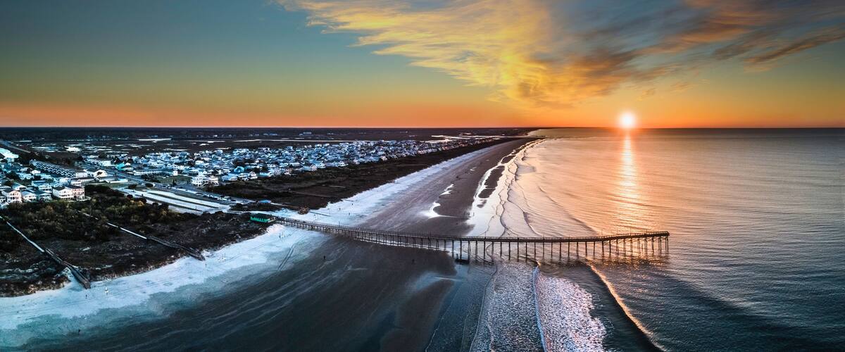 A coastal sunset with a pier extending into the ocean, a beach, and a town in the background.