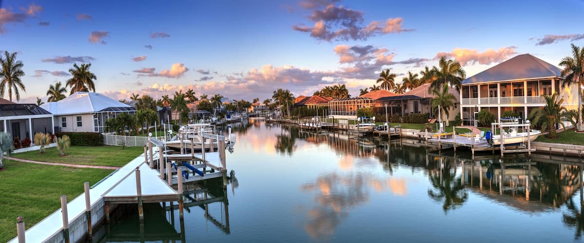 A marina with boats docked, surrounded by houses and palm trees under a clear sky.