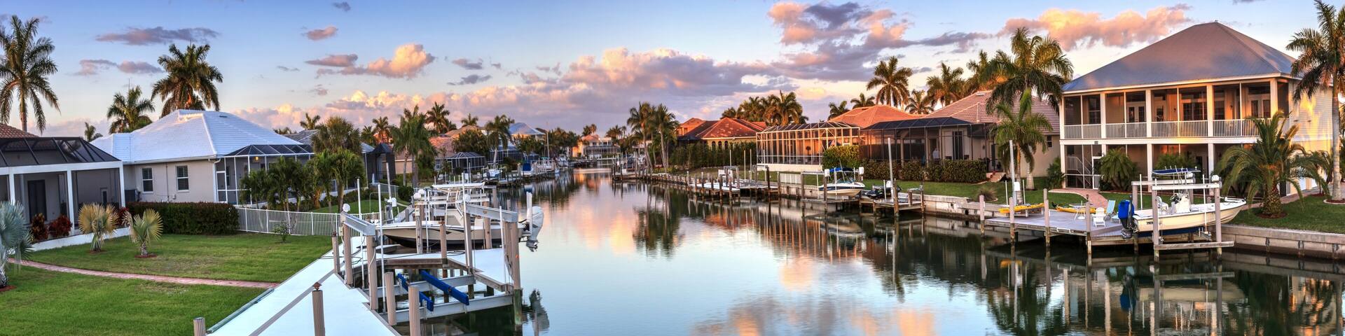 A marina with boats docked, surrounded by houses and palm trees under a clear sky.