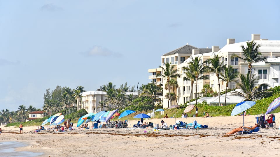 Tourists and locals with umbrellas on the beach with condos in the background at Lantana Beach in South Florida