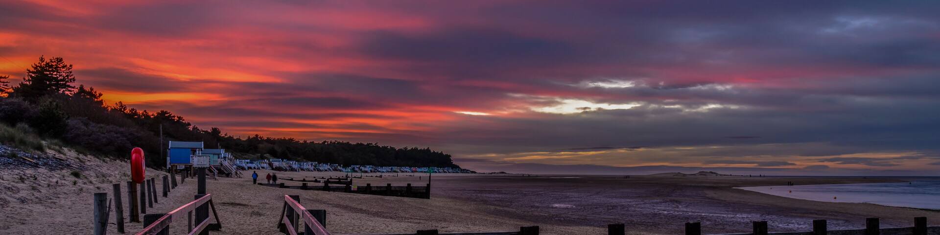 A beach with wooden walkways and beach huts under a colorful sunset sky.