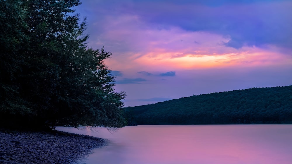 A calm lake with a forested hillside and a colorful sky.