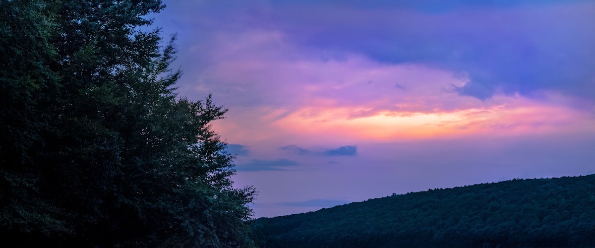 A calm lake with a forested hillside and a colorful sky.