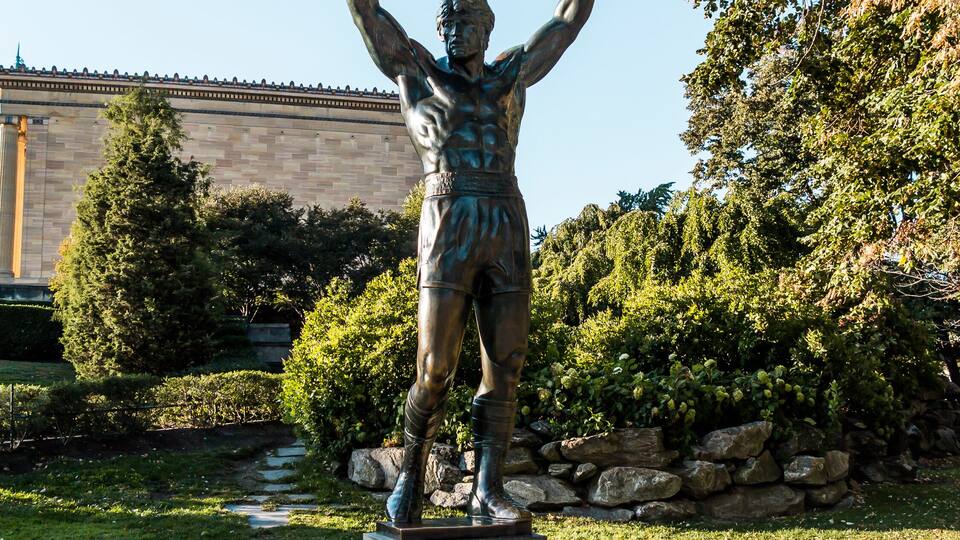Eine bronzene Statue einer muskulösen Figur mit erhobenen Armen, auf einem Sockel in einem Park mit Bäumen und einem Gebäude im Hintergrund.