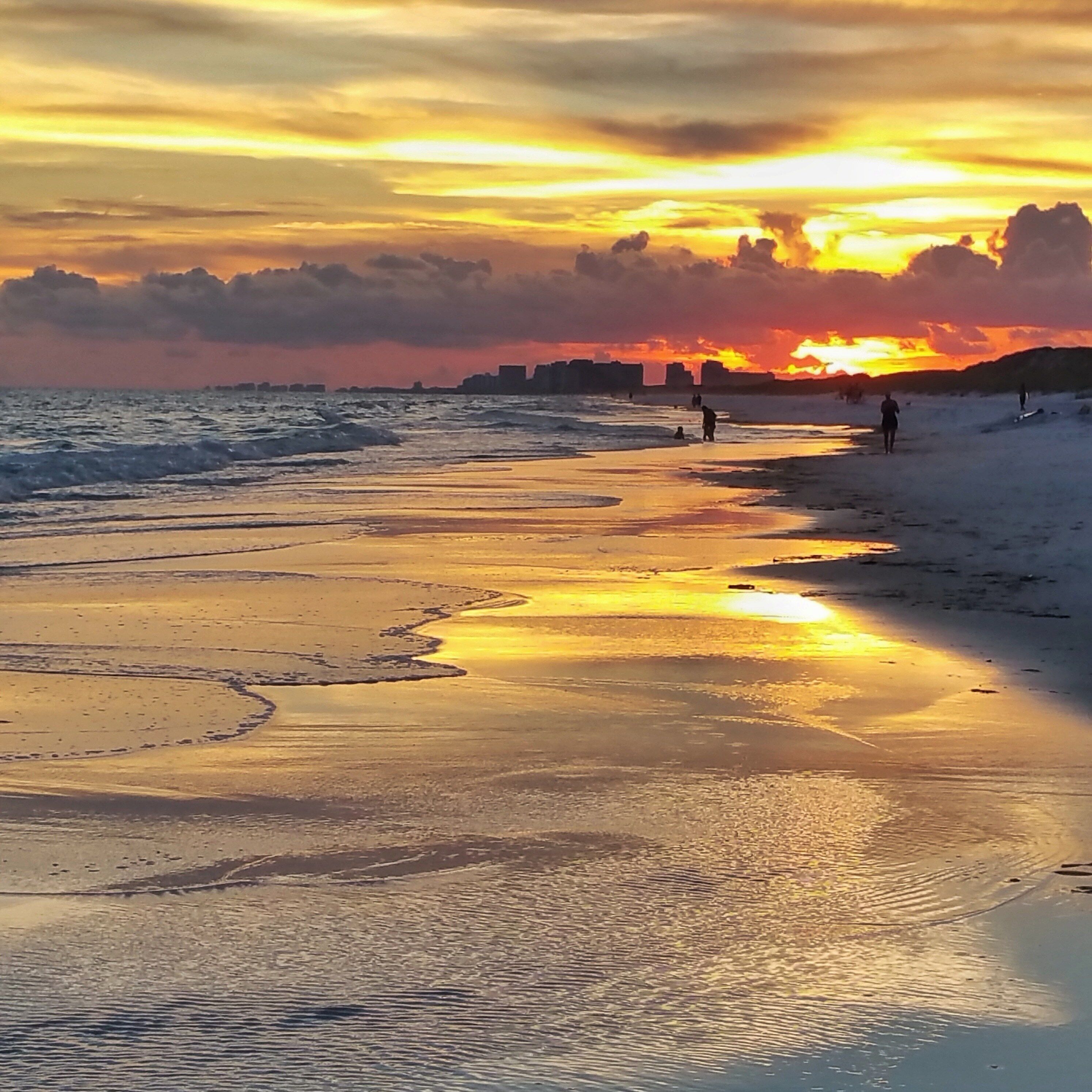 A beach at sunset with waves, people walking, and a city skyline in the background.