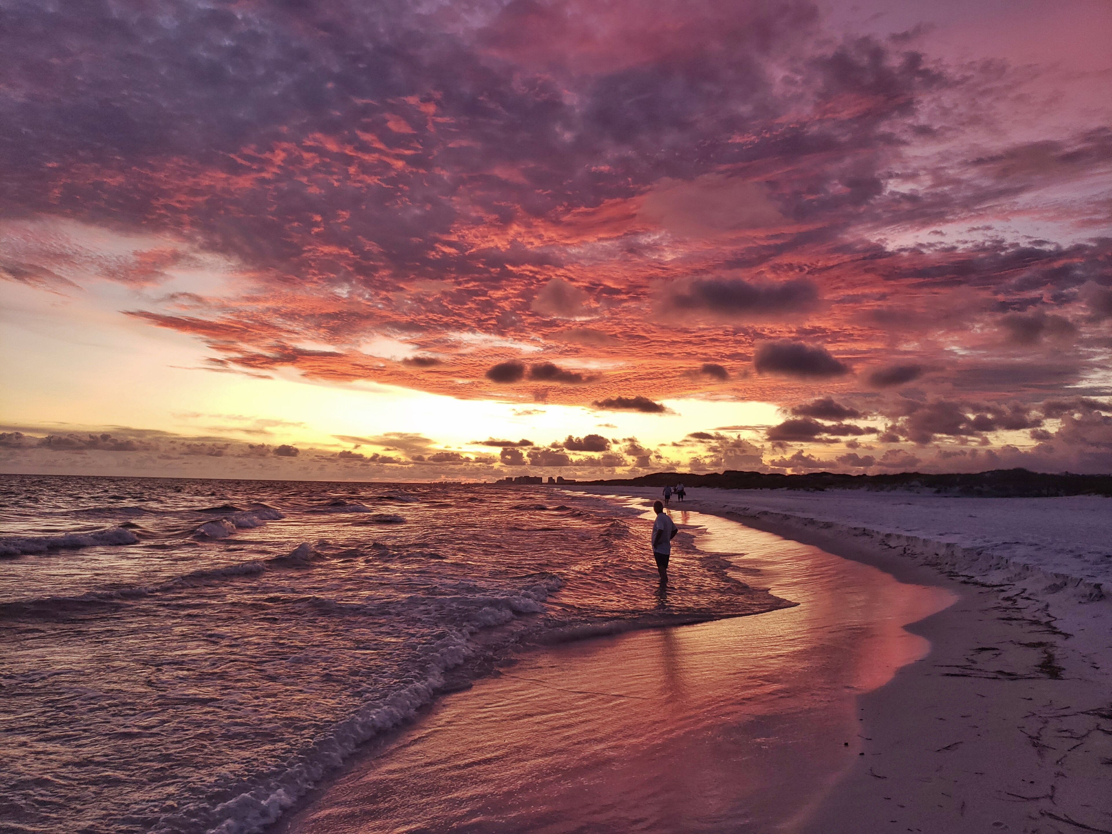 A beach at sunset with waves, a person walking, and a dramatic sky.