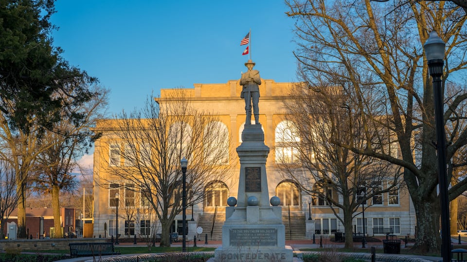 A monument with an American flag, surrounded by trees and benches, in front of a building with arched windows.