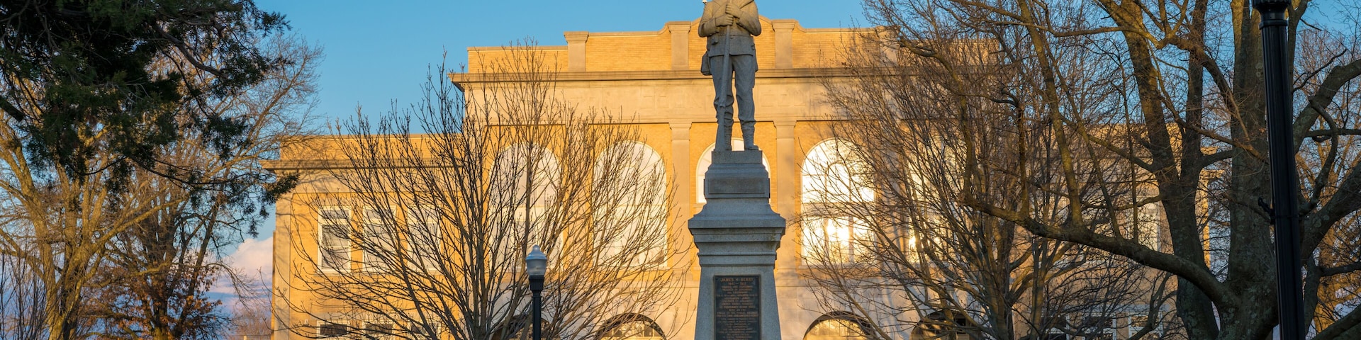 Un monument ornĂ© dâun drapeau amĂ©ricain, entourĂ© dâarbres et de bancs, se dresse devant un bĂątiment dotĂ© de fenĂȘtres en arc de cercle.