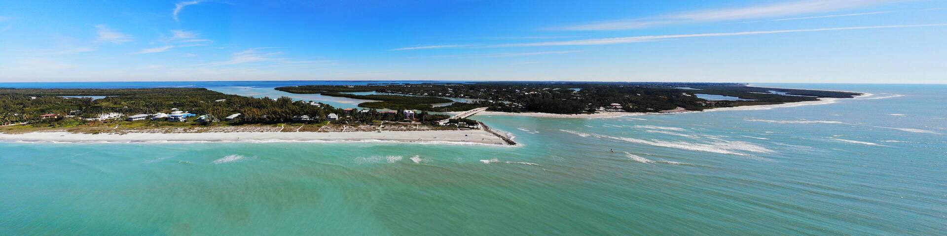 An aerial view of a coastal area with clear turquoise waters, a sandy beach, and a dense cluster of buildings surrounded by greenery.