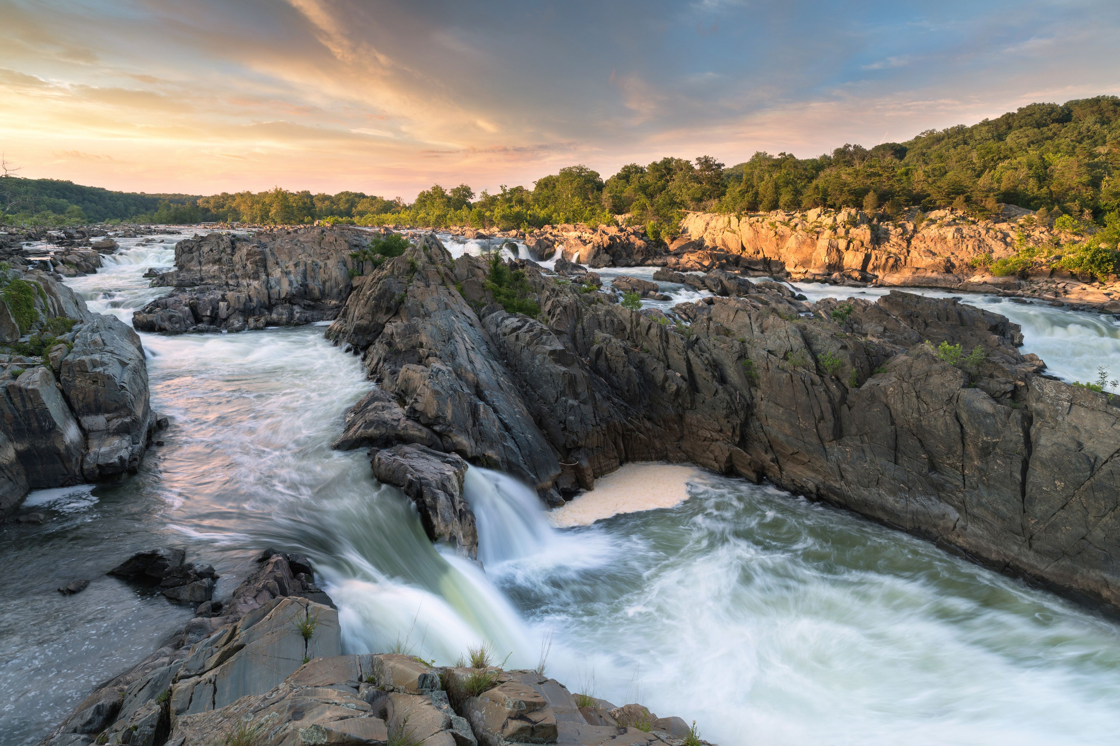 A river flowing through a rocky gorge with waterfalls and a forested hillside in the background.
