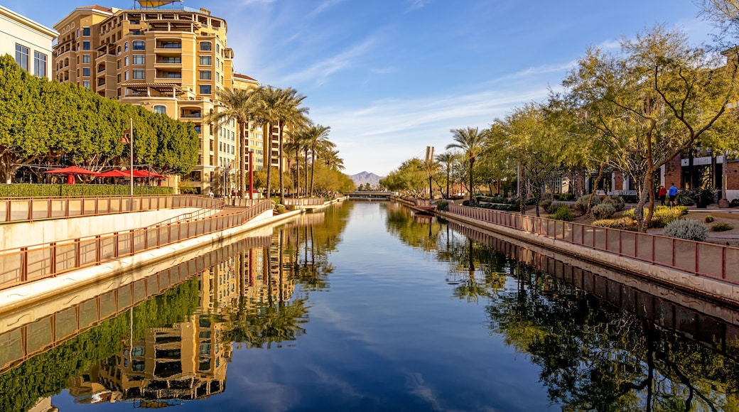 Un canal tranquilo con un puente, bordeado por árboles y edificios, que refleja el cielo y los alrededores.