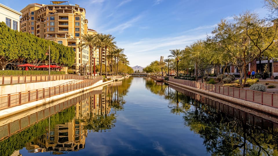 A calm canal with a bridge, lined with trees and buildings, reflecting the sky and surroundings.