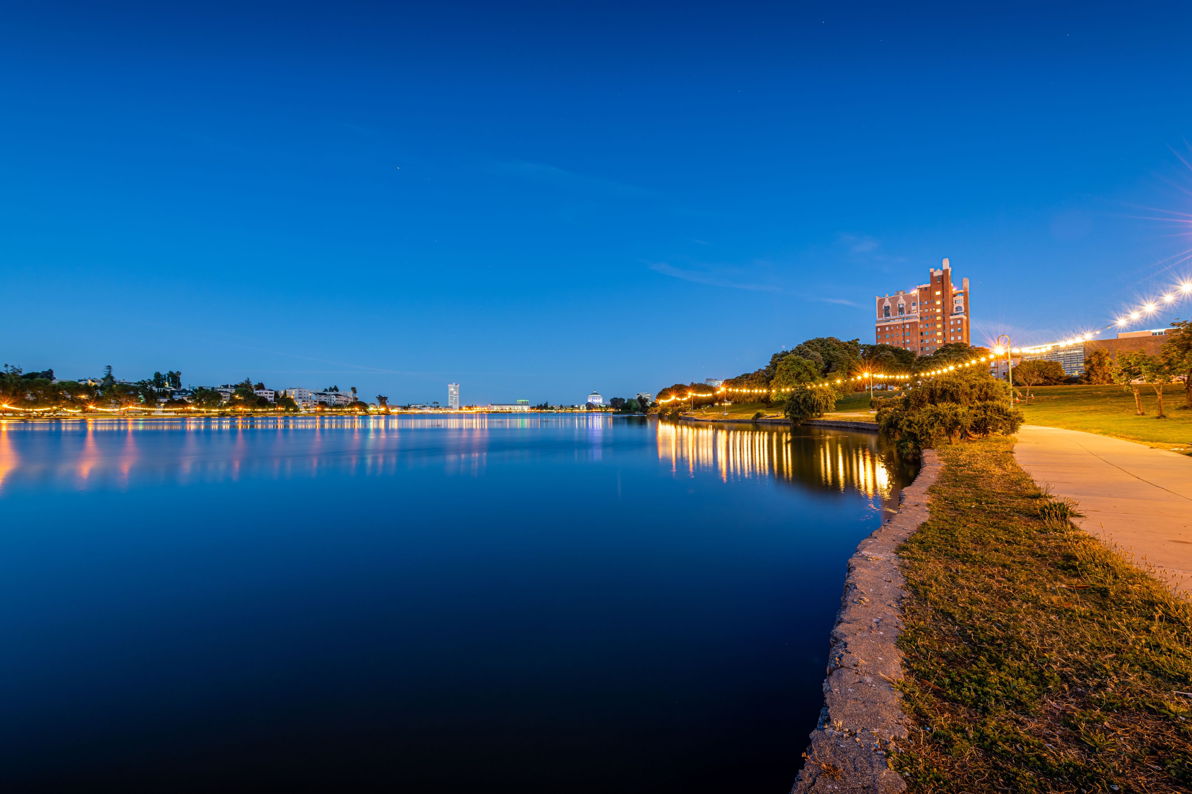 Un lago tranquilo al atardecer con el horizonte de una ciudad al fondo y luces colgantes a lo largo de la orilla.