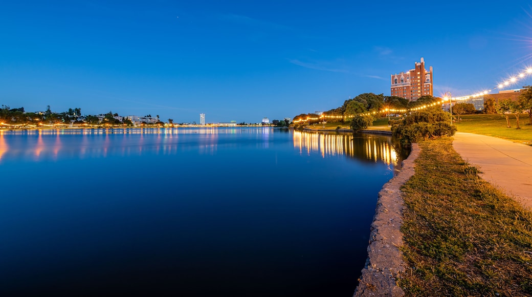 Un lago tranquilo al atardecer con el horizonte de una ciudad al fondo y luces colgantes a lo largo de la orilla.