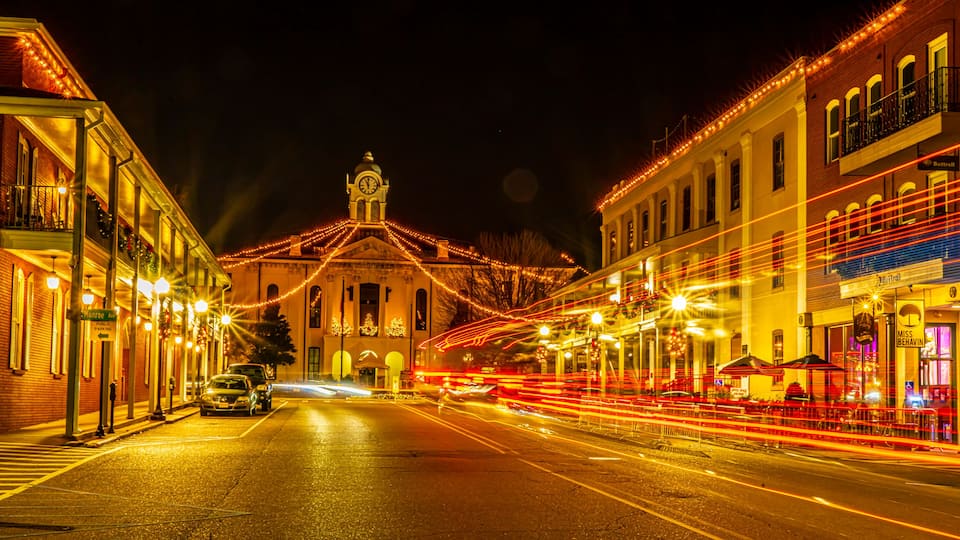 A street at night with buildings on either side, a clock tower, and string lights.