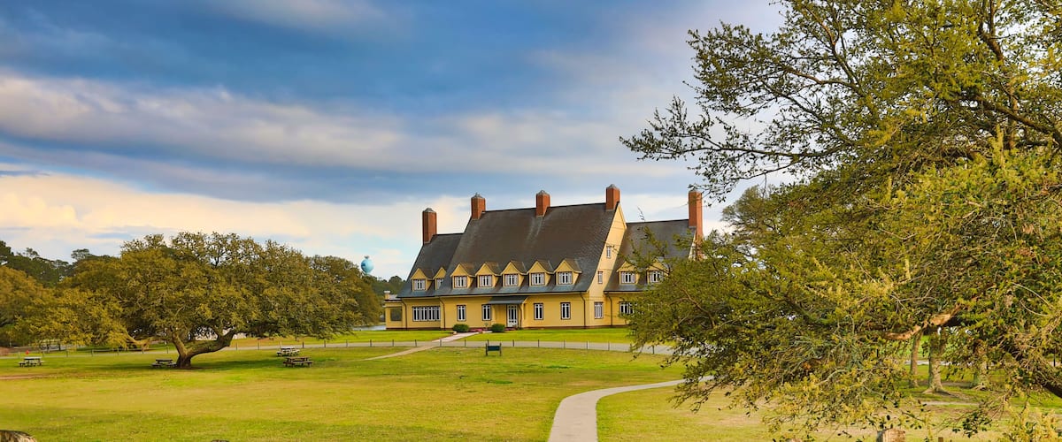 Une promenade en bois mène à une grande maison historique, dotée d’une pelouse verdoyante et bordée d’arbres.