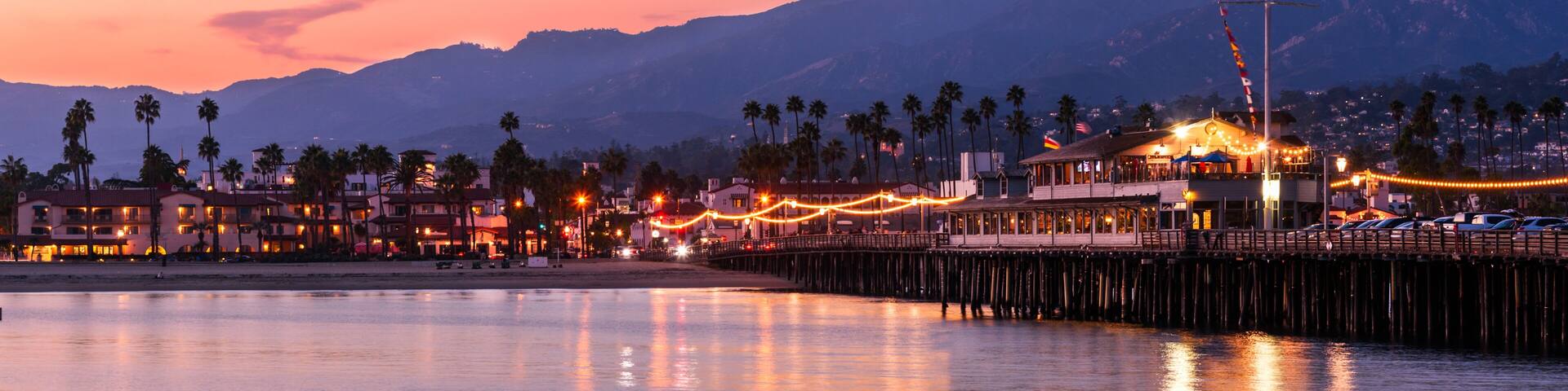 A coastal town at dusk with a pier extending into calm waters, palm trees, and a mountain range in the background.