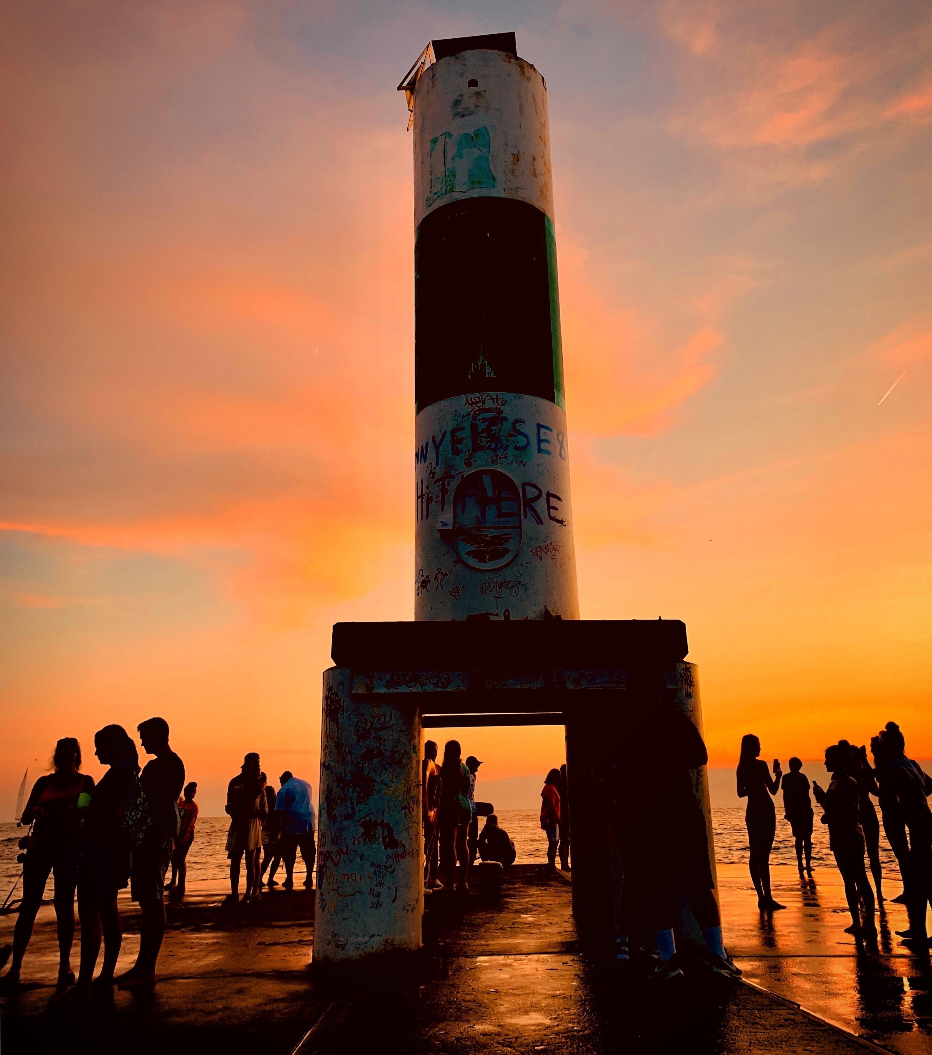 A lighthouse silhouette against a sunset sky, with people gathered around it.