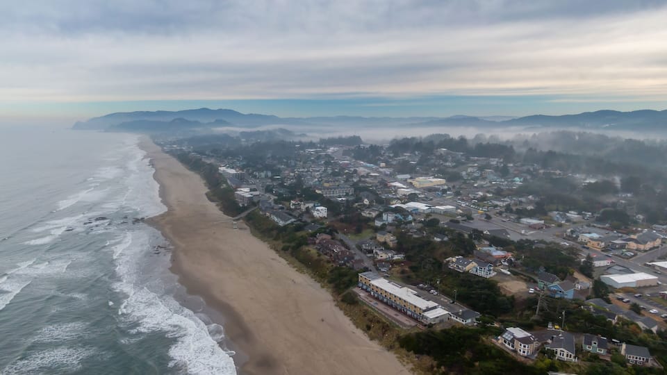 Coastal town of Lincoln City shrouded in fog, waves crashing on the beach, hotels and homes line the shore. Aerial view of a misty morning. Oceanlake, Oregon, USA