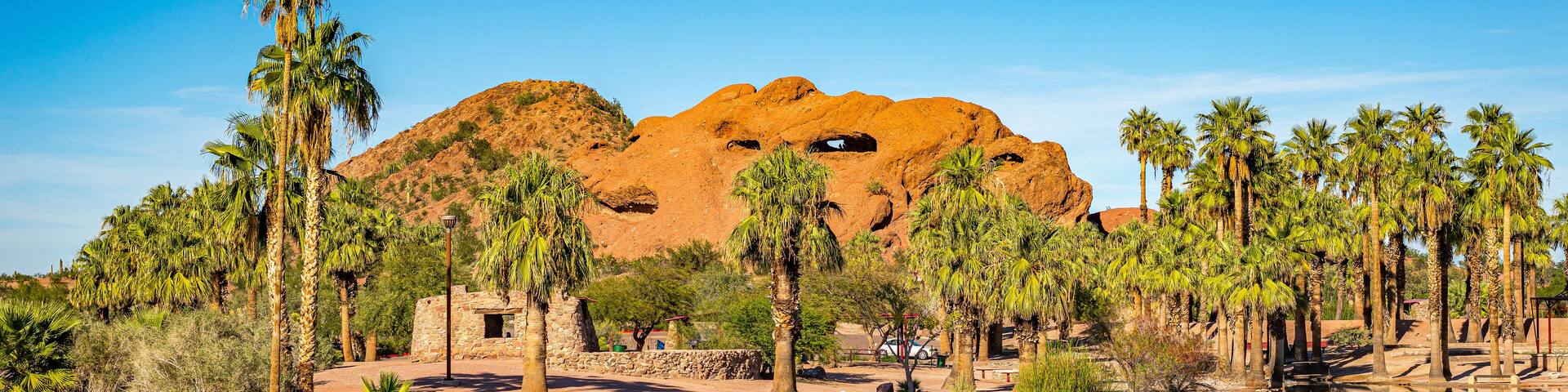 A desert landscape with a large rock formation, palm trees, and a body of water reflecting the scenery.