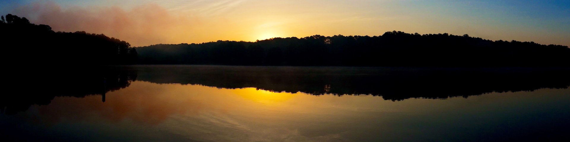 Um lago calmo ao amanhecer, com uma floresta nevoenta ao fundo.