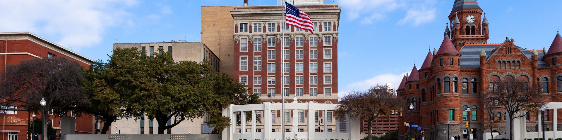 Um edifício histórico com uma torre do relógio, um arranha-céu moderno e um mastro com a bandeira americana.