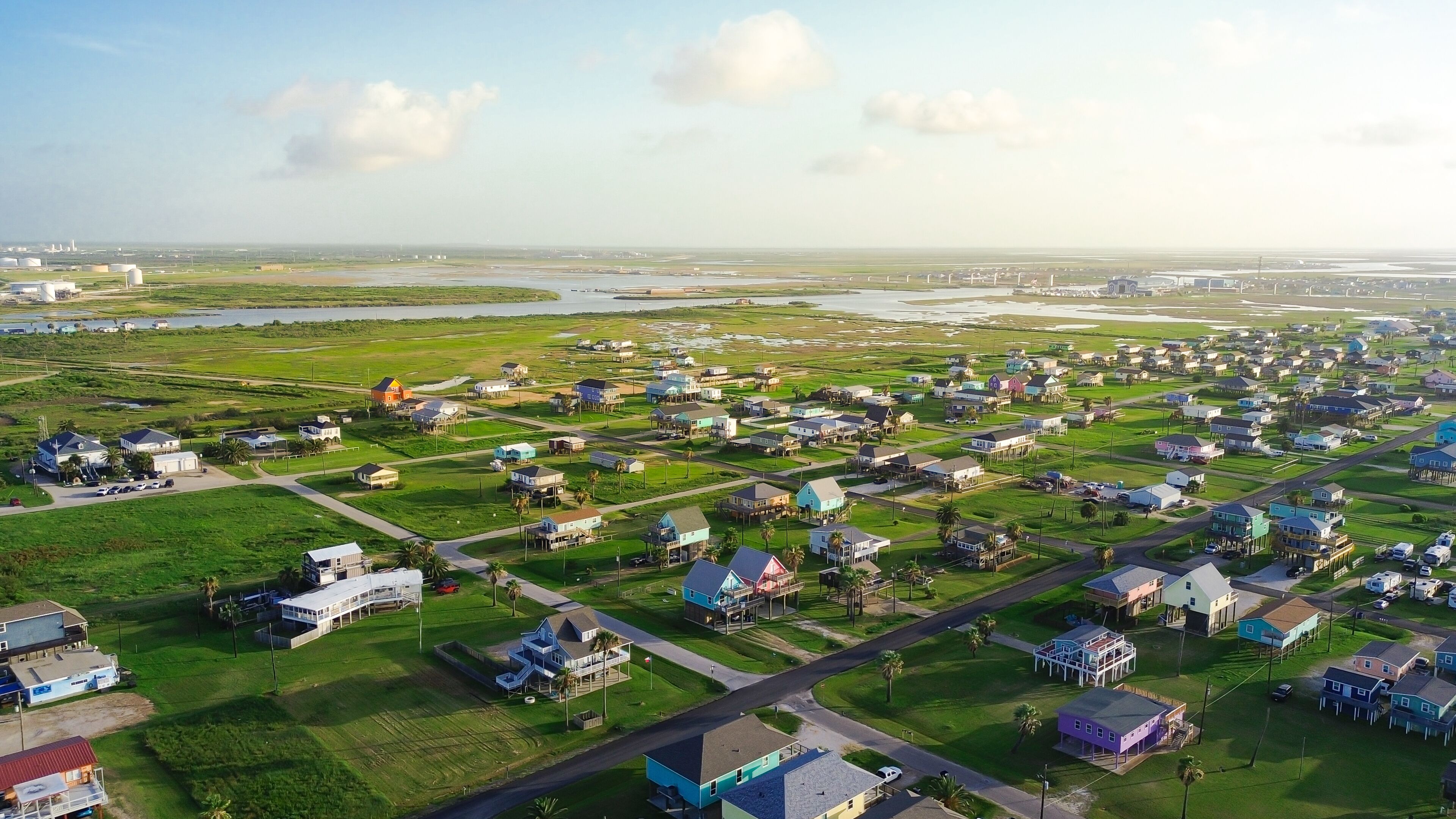 Surfside Beach features colorful stilted homes overlooking marshlands distant industrial infrastructure. Lush green spaces provide vibrant contrast to tidal water systems, Freeport, TX