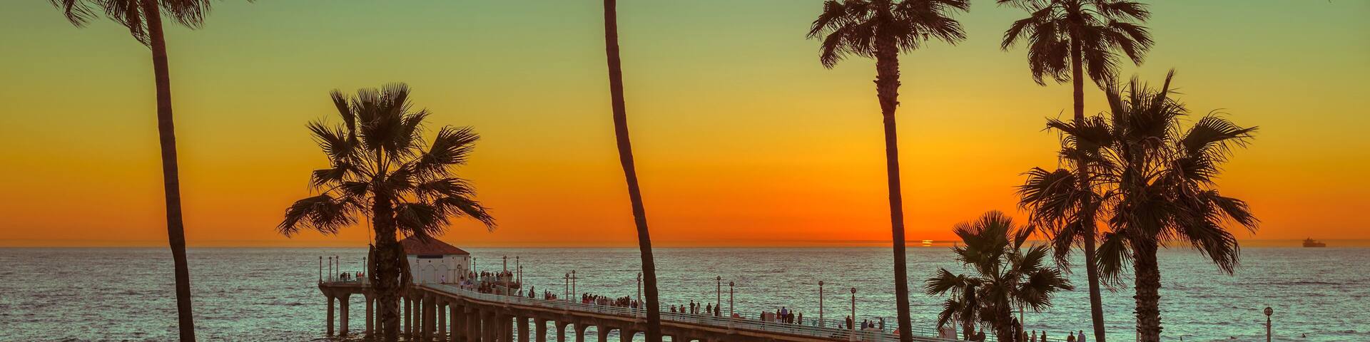 A beach with palm trees, a pier, and the ocean at sunset.